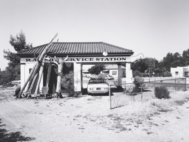 Old black and white photo of a service station with dried cacti resting upon it and a convertible parked underneath the station