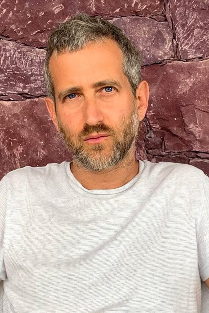 Portrait of artist Yoshua Okón, White shirt in front of a rocky wall background.